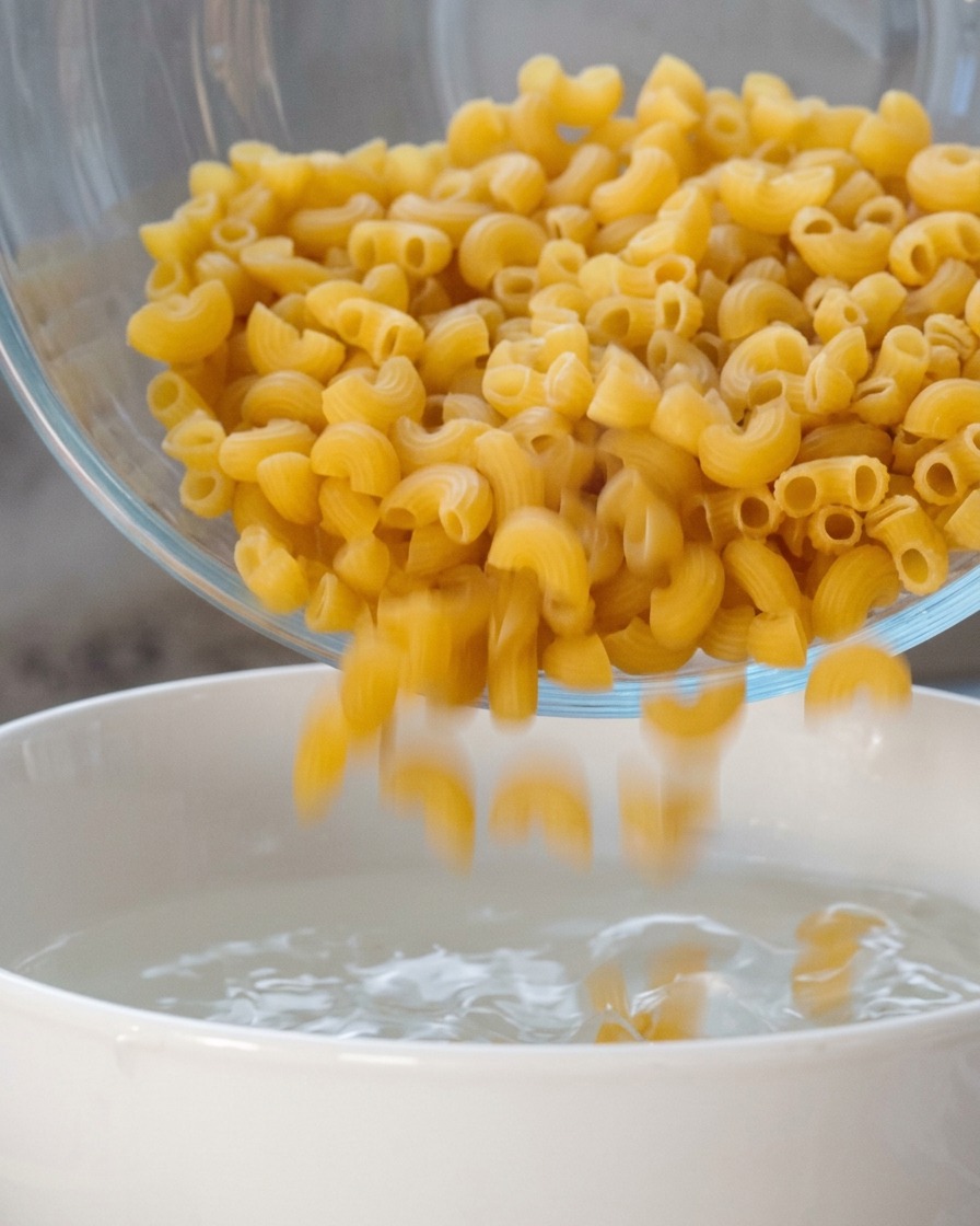 ボウルに入った乾燥スパイラルマカロニを、沸騰したお湯が入った白い鍋に入れている様子。