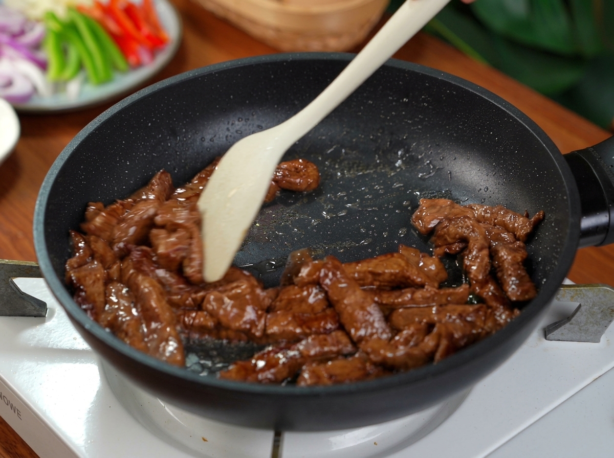 シリコンヘラを使い、黒いフライパンで下味をつけた牛肉の細切りを素早く炒めている様子。