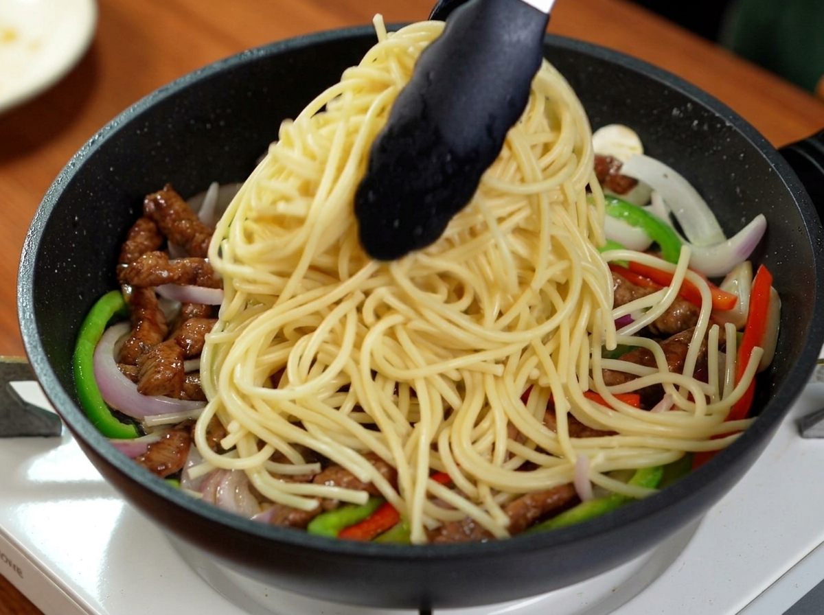 牛肉やピーマンが入ったフライパンに、茹で上がった大量のスパゲッティをトングで投入している様子。