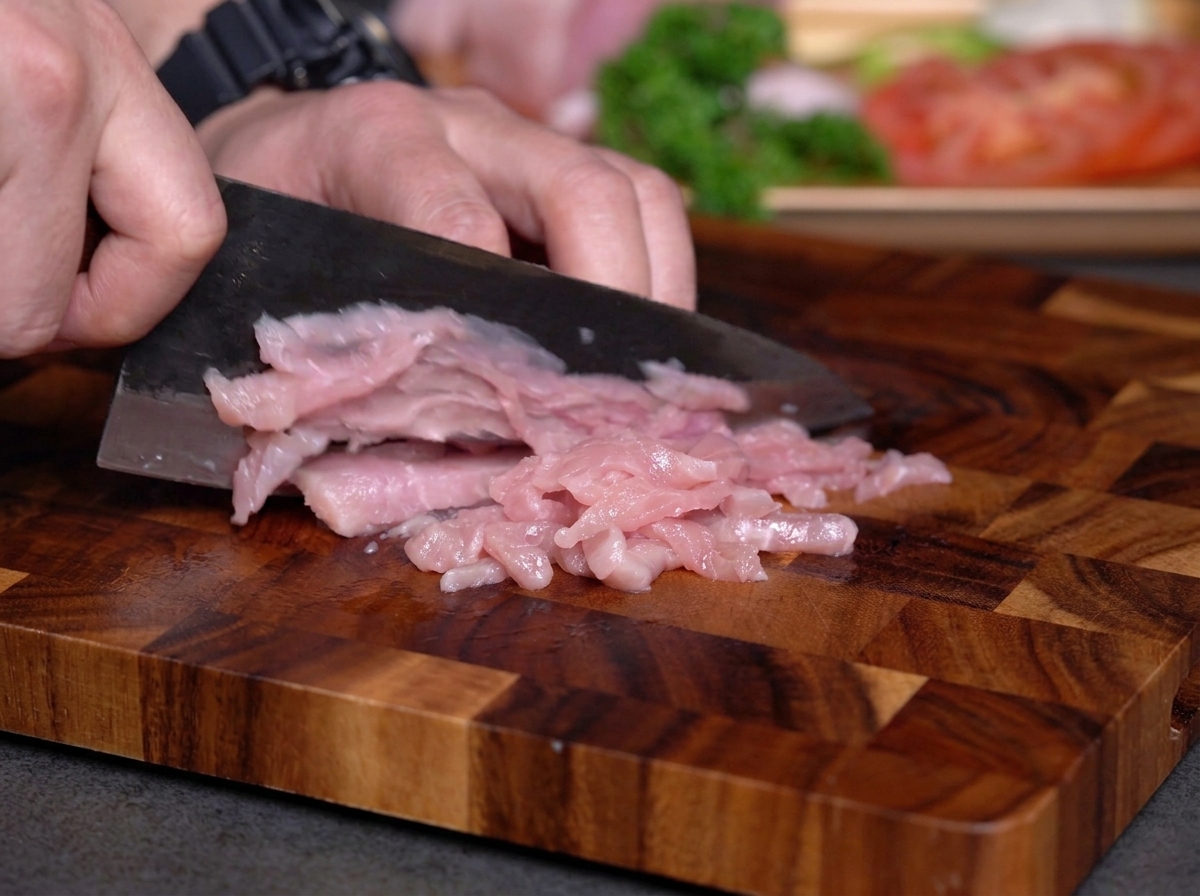 木製のまな板の上で、黒い包丁を使って生の赤身肉を細切りにしている様子。
