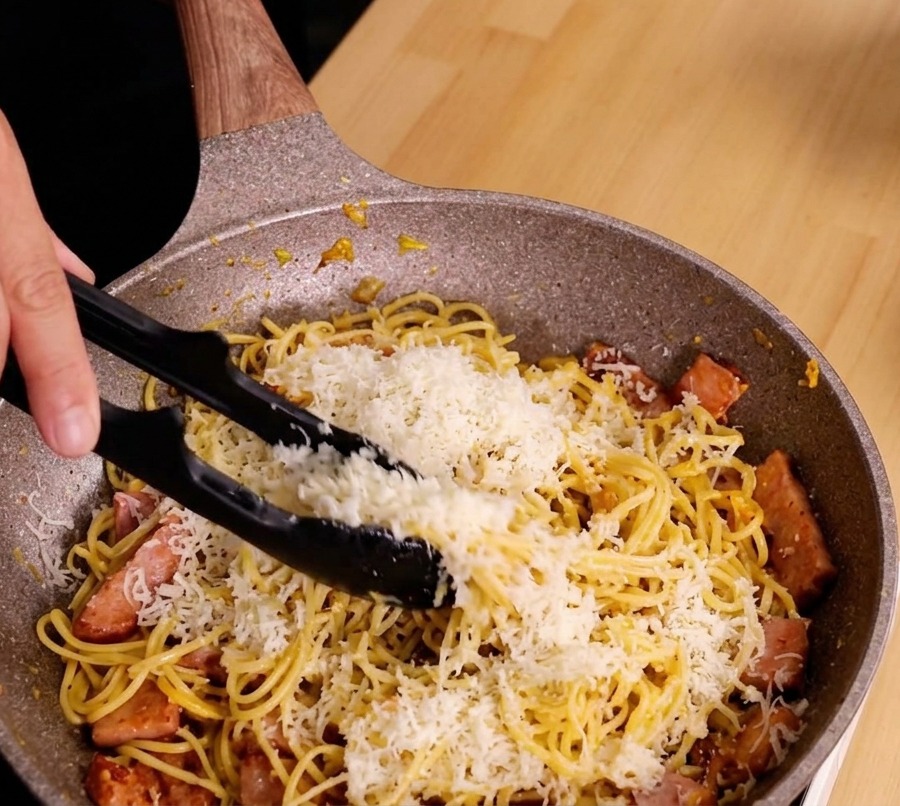 トングを使って、パスタ、チーズ、ベーコンを力強く混ぜ、クリーミーなソースを作っている様子。