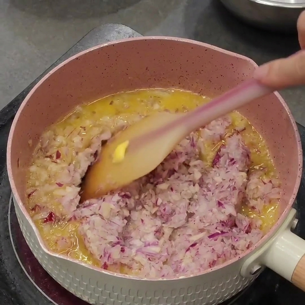 ピンクの鍋の中で、みじん切りの紫玉ねぎとニンニクがバターで炒められている様子。