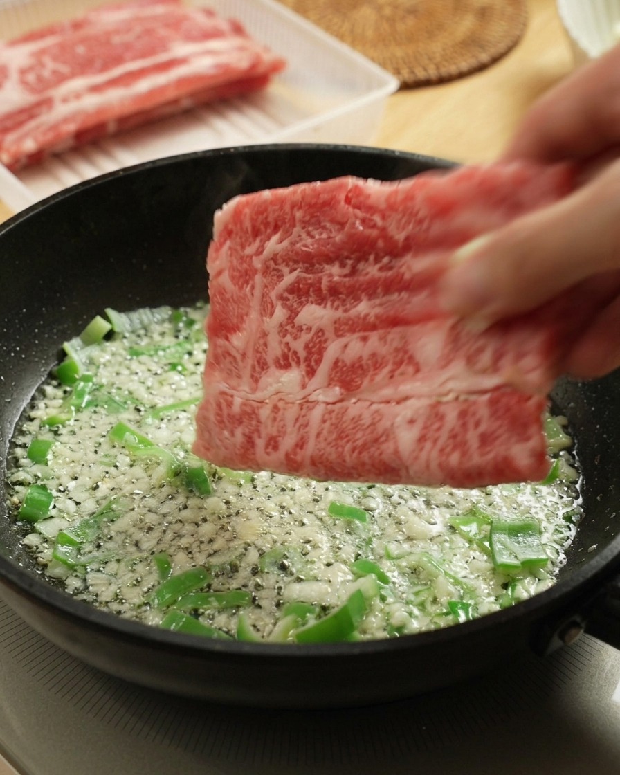 にんにくとねぎが入ったフライパンに、生の牛薄切り肉を投入する様子。