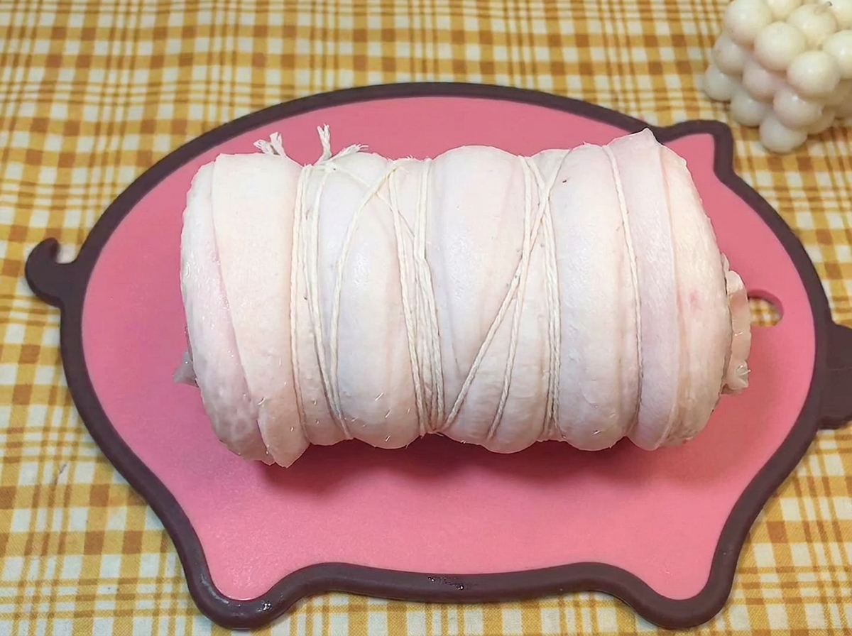 ピンクのボードの上で、料理用の白いタコ糸でしっかり縛られた円柱状の豚バラ肉。