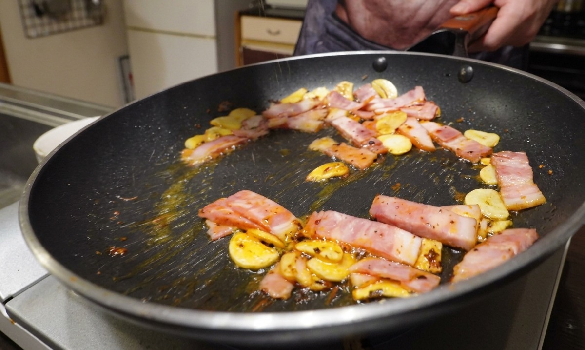 にんにくとチリフレークが入ったフライパンにベーコンを加え、黒こしょうを振っている様子。