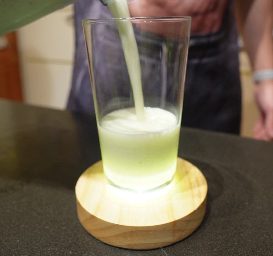 ミキサーで撹拌された薄緑色のクリーミーなドリンクを、背の高いグラスに注いでいる様子。