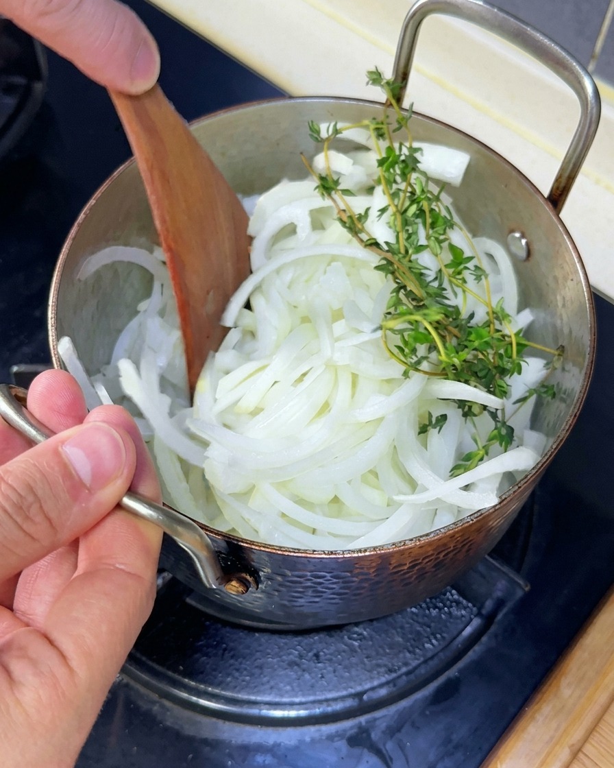 鍋の中で玉ねぎとタイムの枝を木べらで混ぜ合わせている様子。