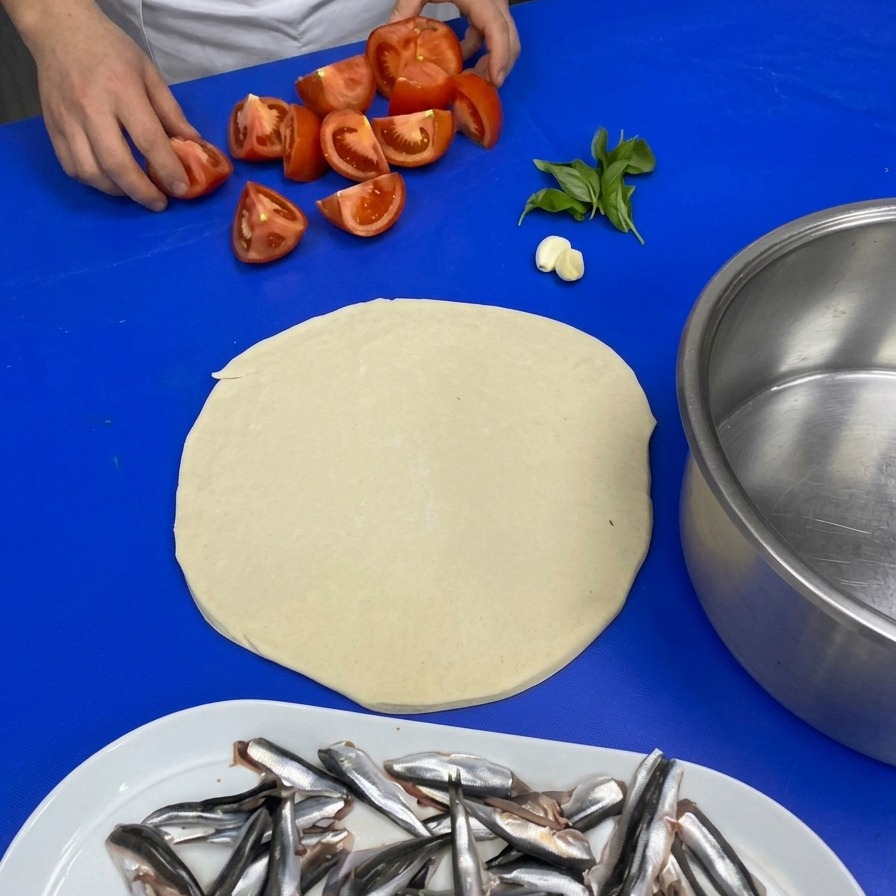 青い作業台の上にある4等分に切ったトマト、にんにく、フレッシュバジルの葉を手に取るシェフの手。