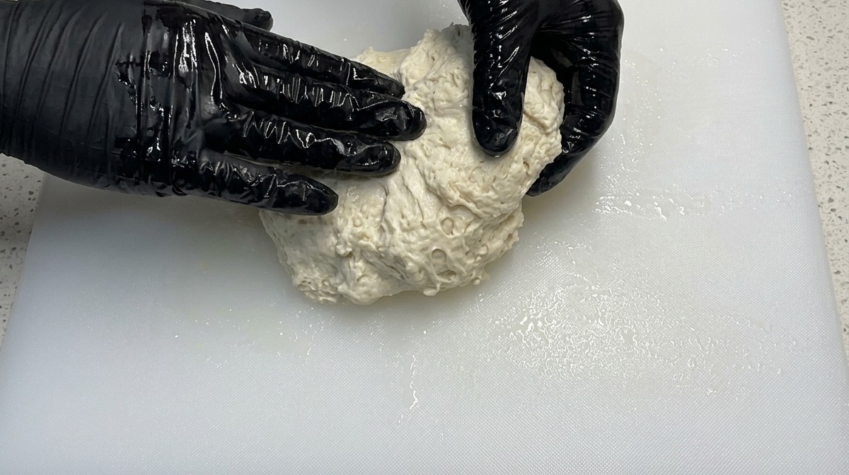 黒い手袋をした手で、白いまな板の上の柔らかいピザ生地をこねている様子。