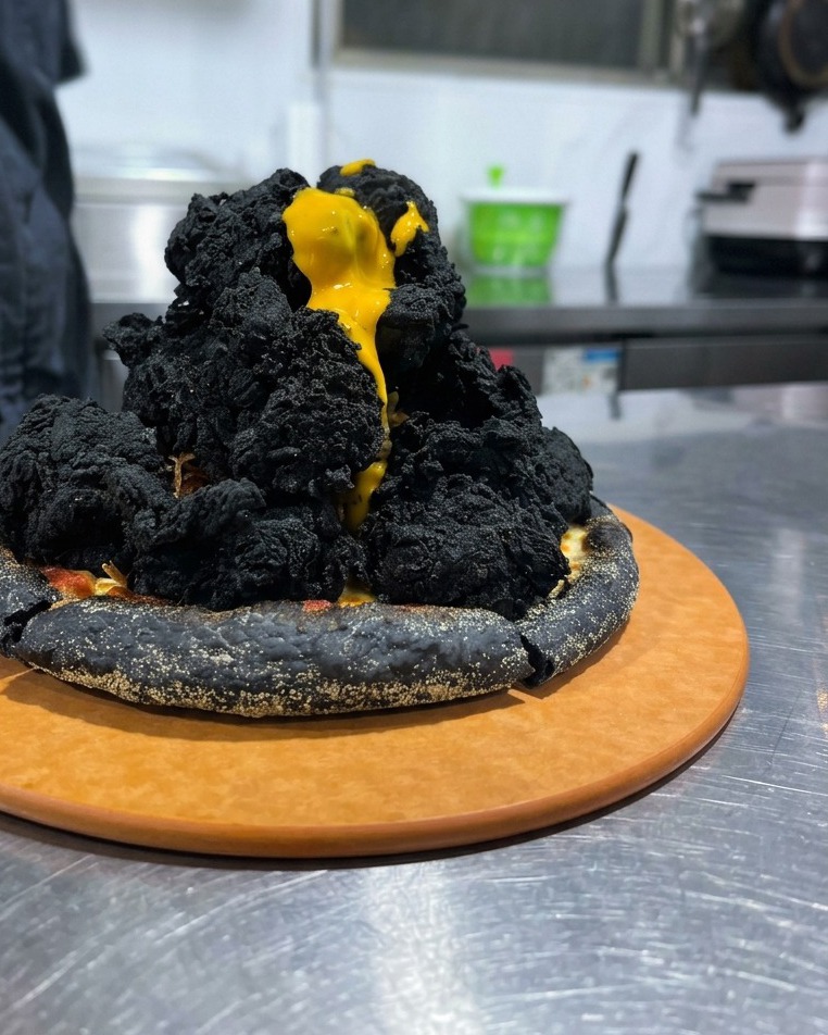 積み上げられた黒いチキンの頂上から、鮮やかな黄色のチーズソースが溢れ出している迫力あるシーン。