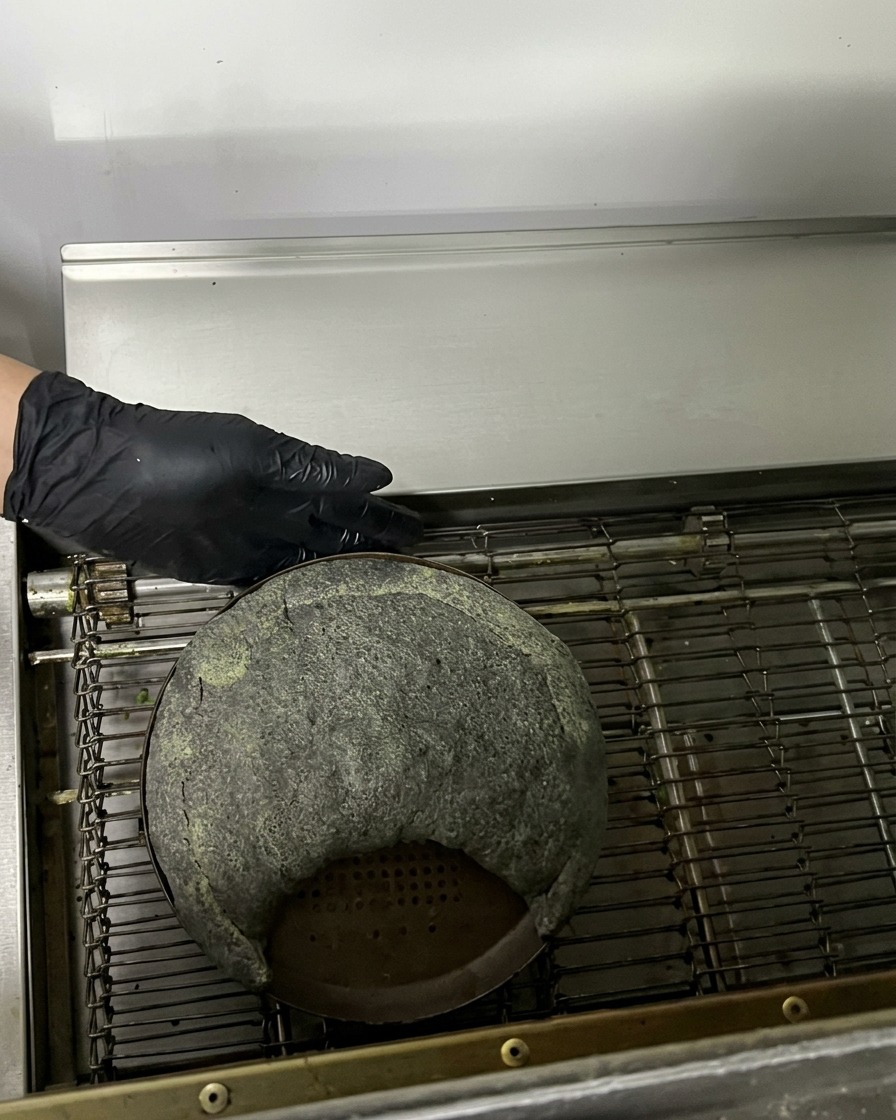 金属製の天板に乗った焼く前の黒いカルツォーネが、コンベアオーブンに入っていく様子。