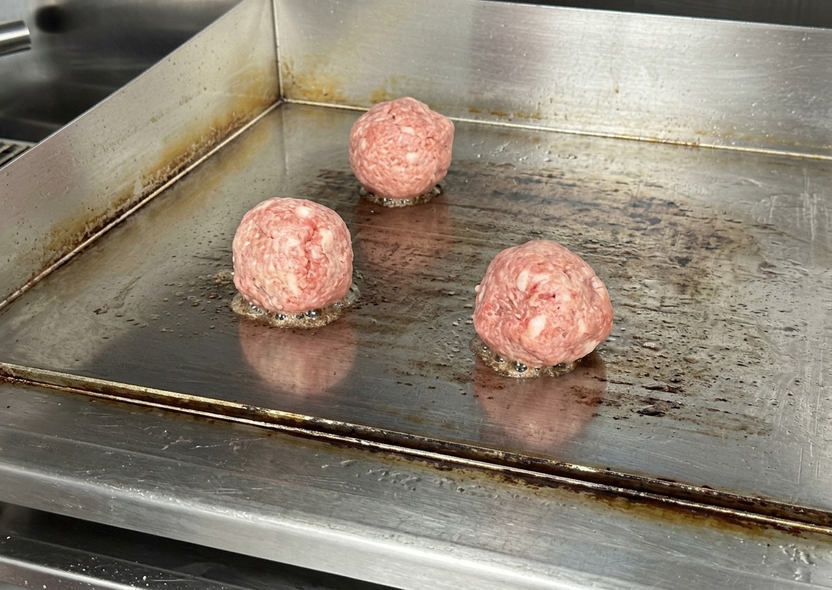 熱したステンレスの鉄板の上に、丸く成形された3つの生の牛ひき肉が並び、底からジューと音がしている様子