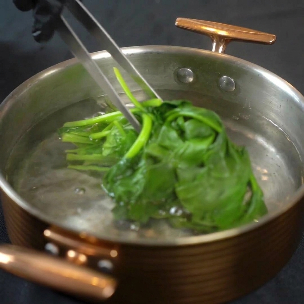 茹でた鮮やかな緑色のほうれん草を、トングで持ち上げて鍋から冷水のボウルに移す様子。