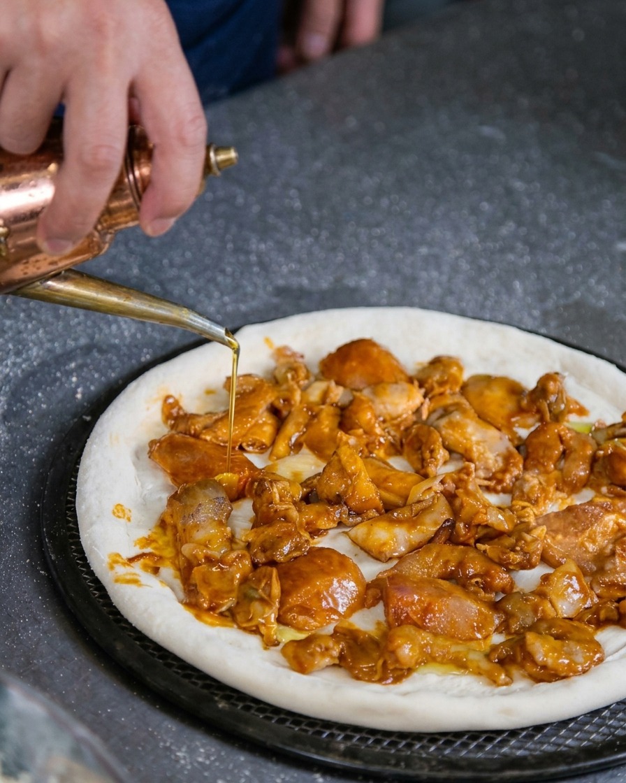 マリネした魚介類がたっぷりのった焼く前のピザに、銅製のオイルディスペンサーを使ってオリーブオイルを優しく回しかけている様子。