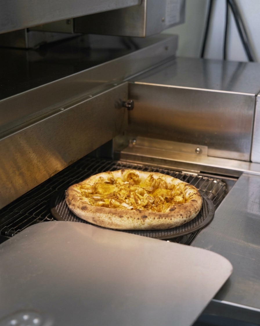 業務用のコンベアオーブンの中で、生地が膨らんで焼き色がつきながら進んでいくシーフードピザ。
