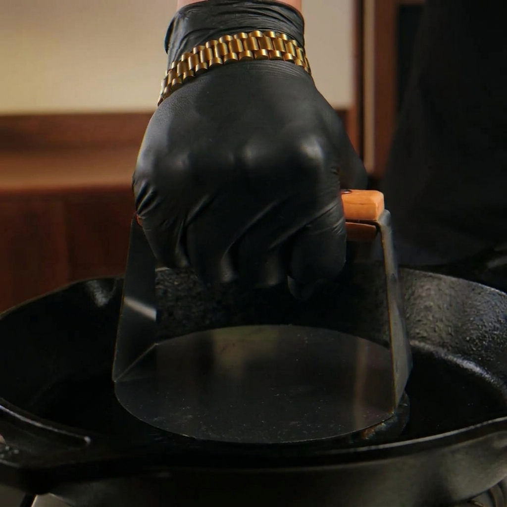 手袋をした手が、金属製のミートプレスを使って熱いスキレットの上で肉のボールを平らに押し潰している様子。
