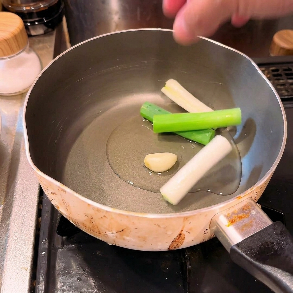 冷たい油が入った鍋に、ぶつ切りにした長ねぎとにんにくを入れている様子。