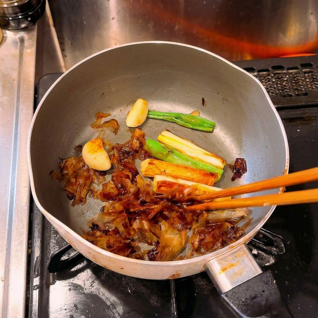 弱火で加熱され、濃い茶色に色づいたねぎ、にんにく、鰹節を箸で混ぜている様子。