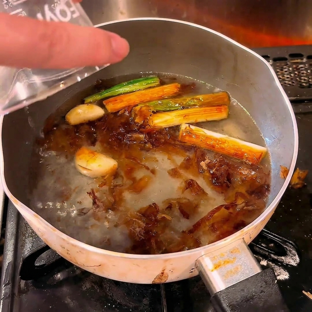 こんがりと焼けた香味野菜が入った熱い鍋に、計量カップから水を注いでいる様子。