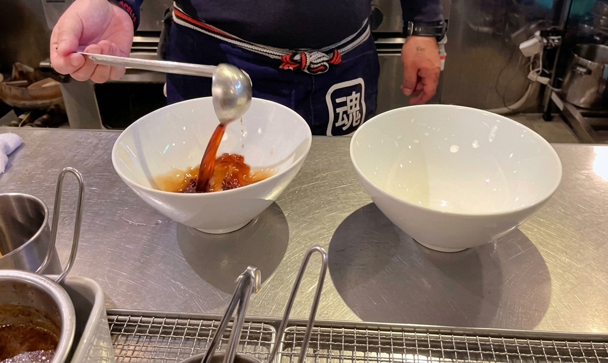 白いラーメン鉢に濃い色の醤油タレをレードルで注ぐ様子