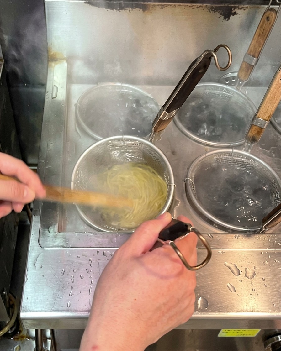 沸騰したお湯の中で、テボ(振りザル)に入った生麺を茹でている様子。