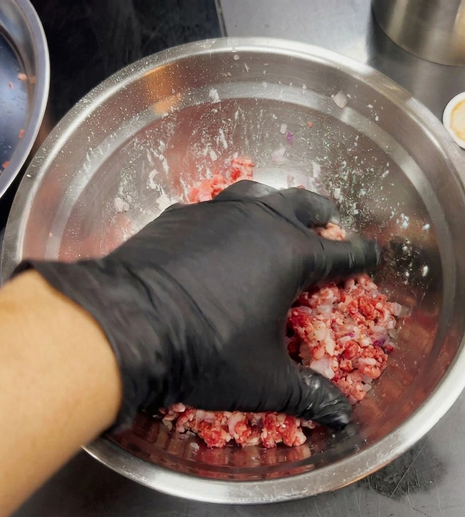 黒い手袋をはめた手で、ひき肉と赤玉ねぎをボウルの中で軽く混ぜている様子。