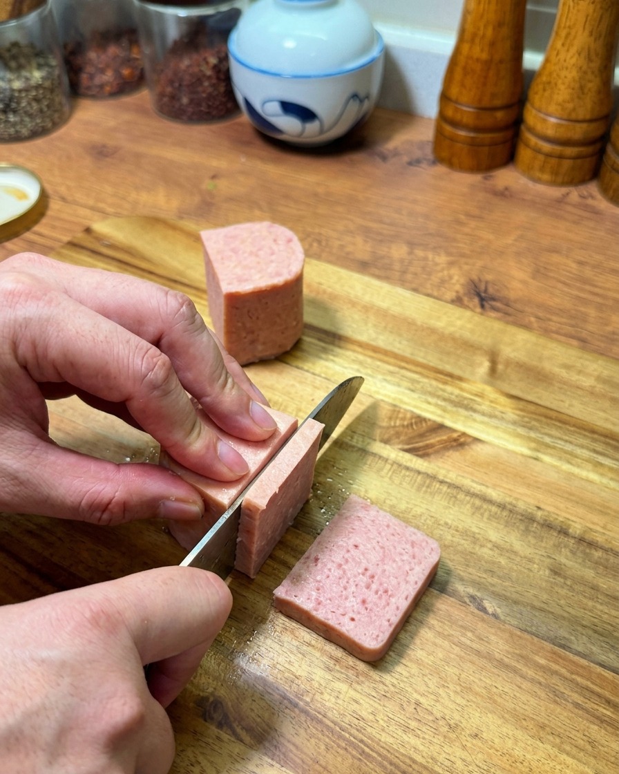 ピンク色のランチョンミートをまな板の上で厚めの長方形にスライスする様子。