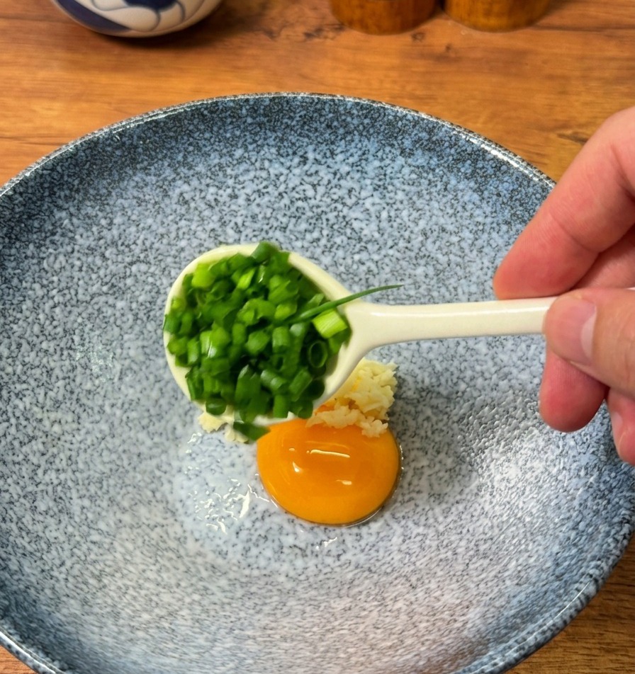 器の中に、刻んだねぎ、おろしにんにく、粉末スープ、そして生卵の卵黄を加える様子。