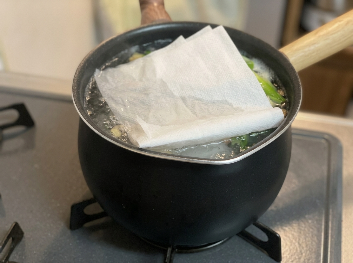 コンロに置かれた黒い鍋の中で、沸騰する煮汁の上に折りたたんだ白いキッチンペーパーがかぶせられている様子。