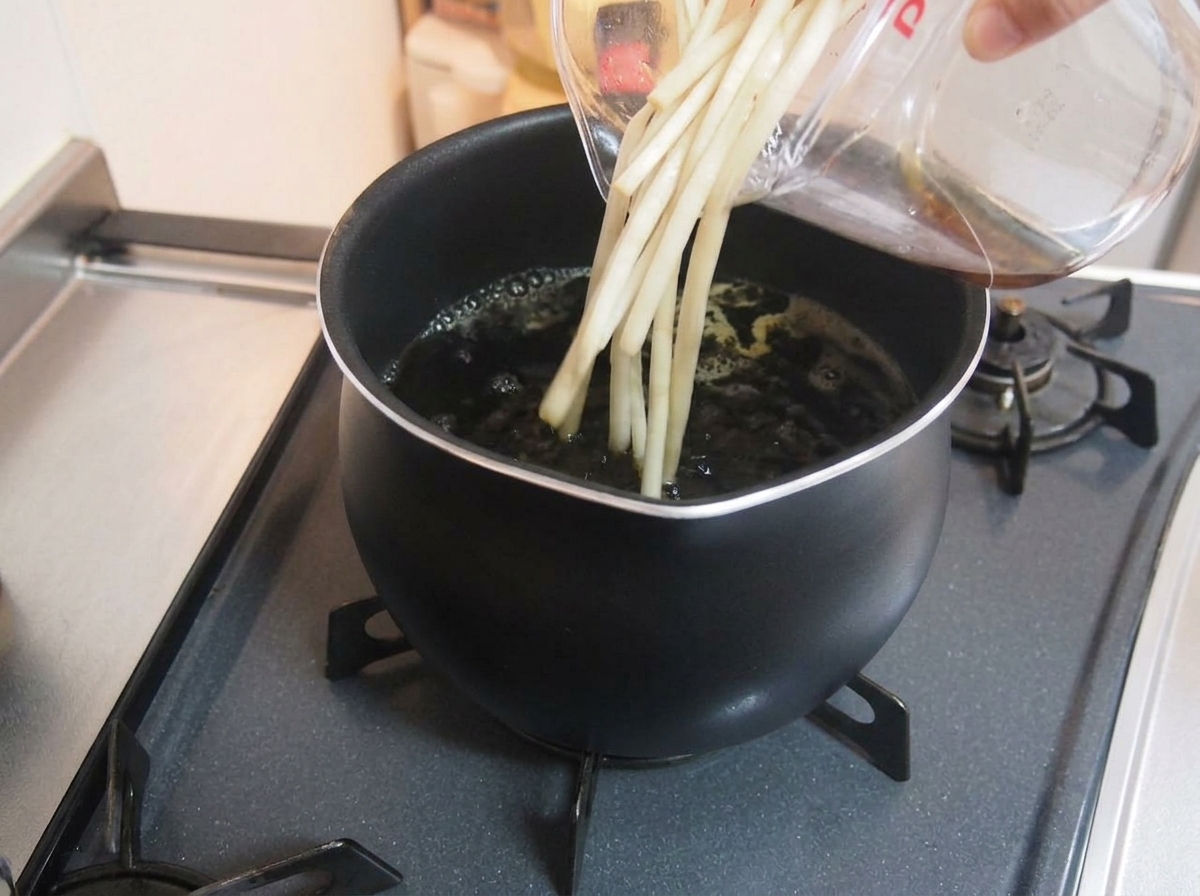 透明な計量カップから、煮立っている濃い色の煮汁の鍋に、液体を注いでいる様子。