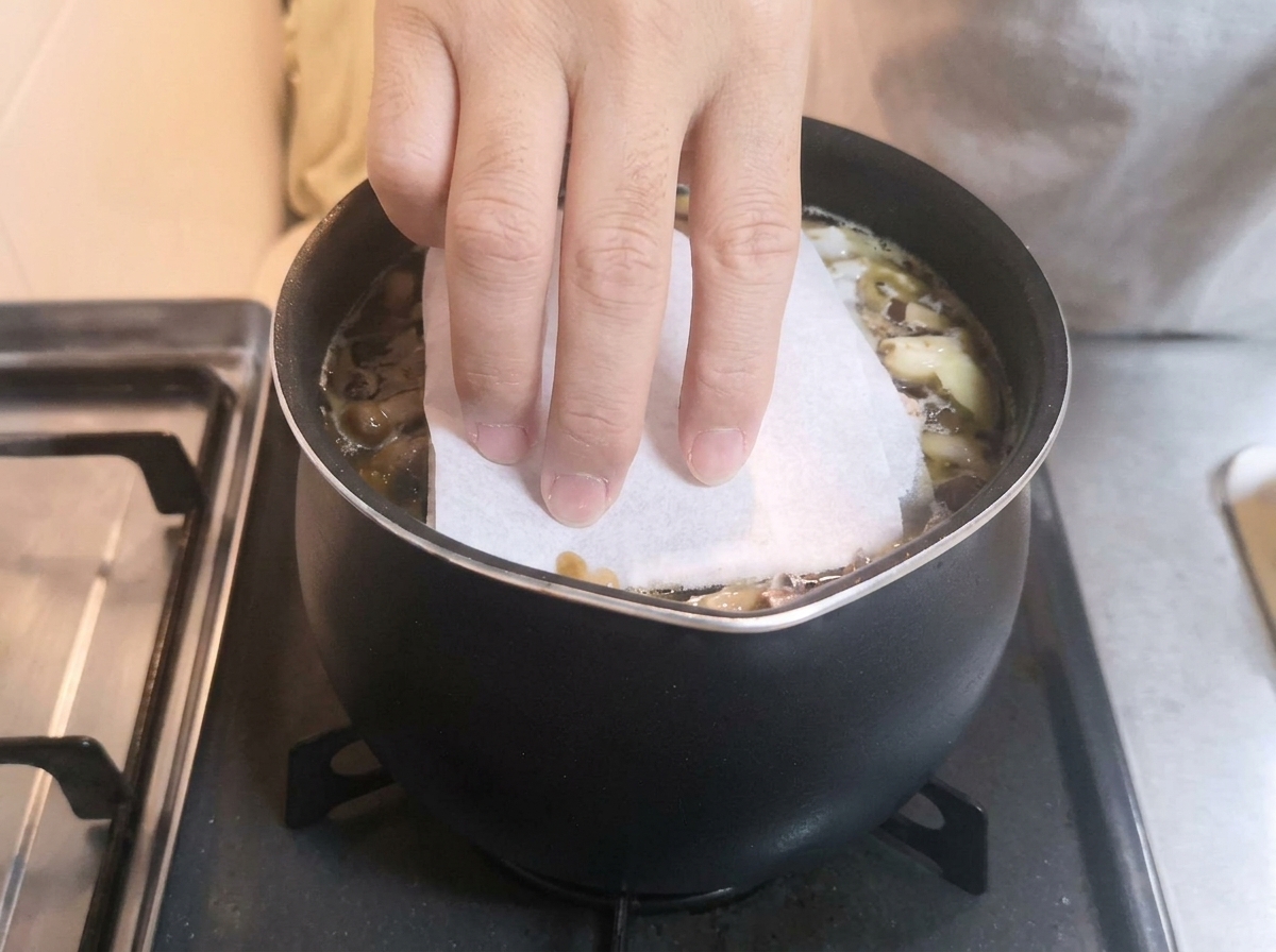 濃い色の煮汁が入った鍋の上に、キッチンペーパーで落とし蓋をしている手。