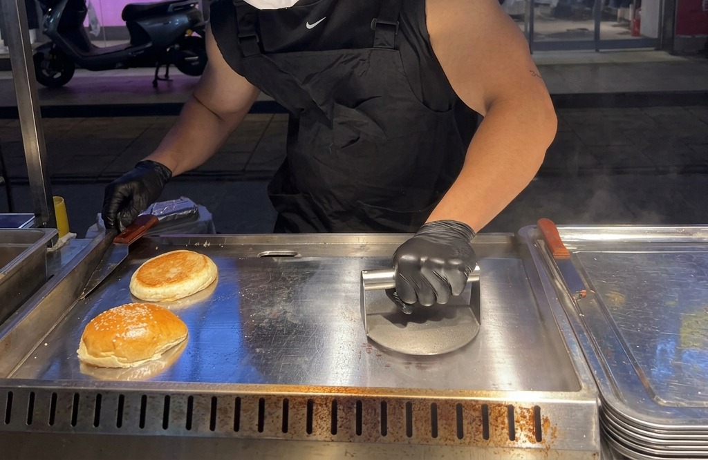 重いバーガープレスを使って、肉の塊を鉄板の上で平らに押し潰している様子。