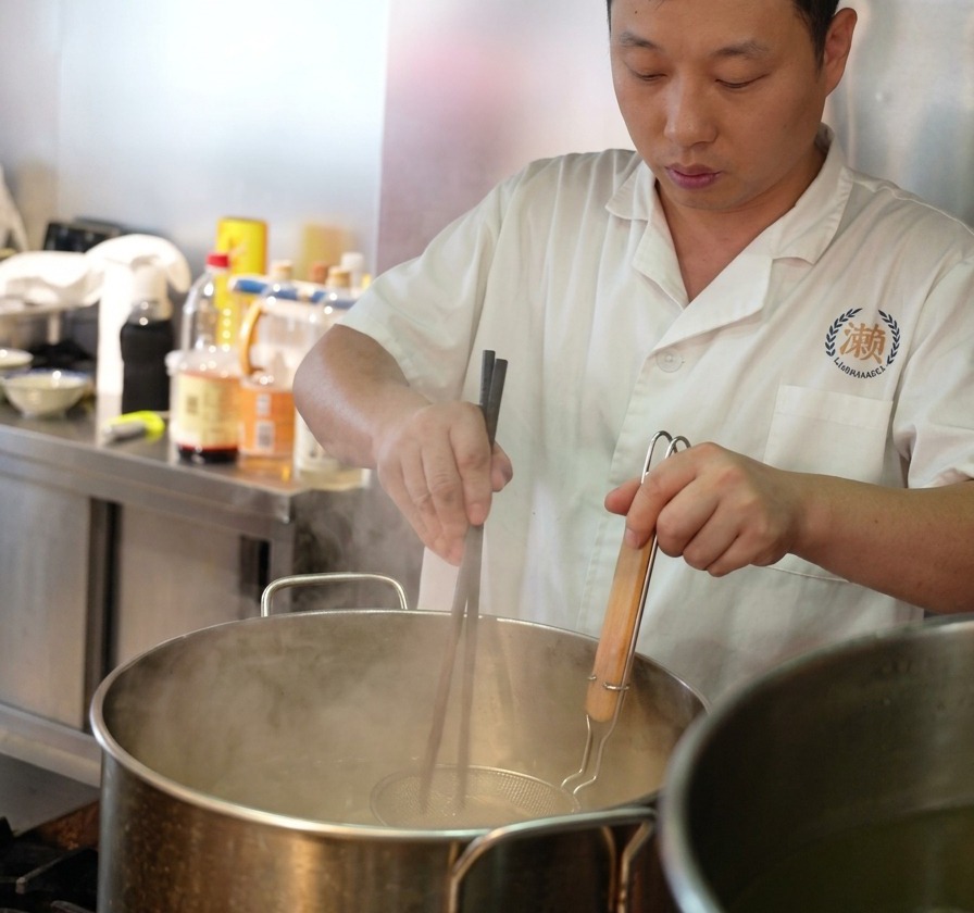 大きな鍋の沸騰したお湯の中で、網を使って生麺を茹で、菜箸で優しく混ぜている様子。