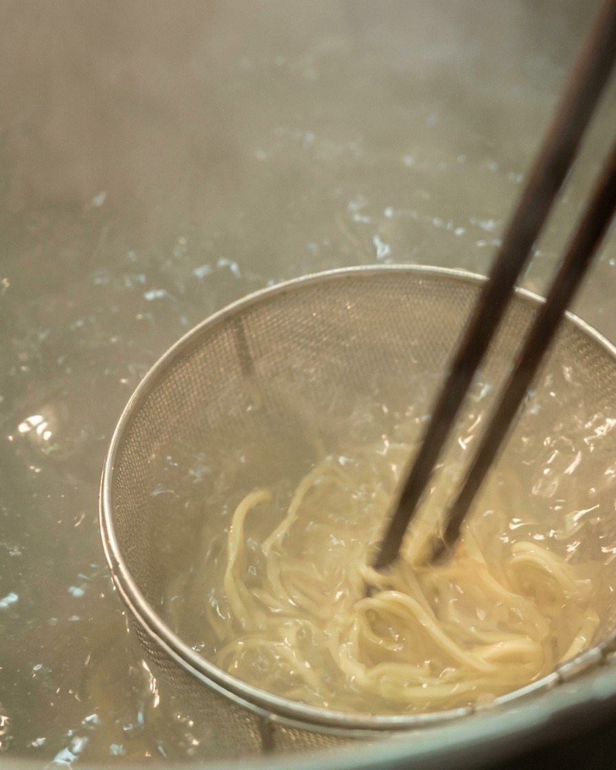 沸騰したお湯の中で、網に入った薄い色のラーメンの麺を菜箸で混ぜているクローズアップ。