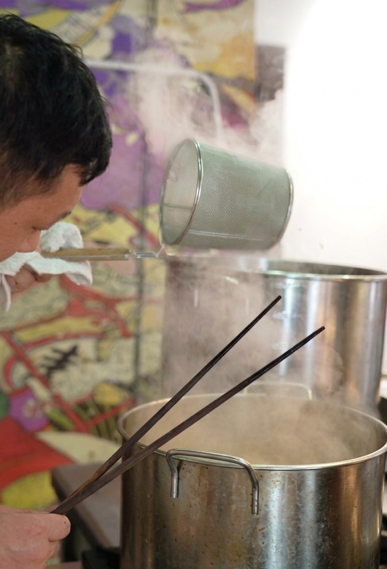 蒸気が上がる鍋の上で、円筒形の網を振り、麺の水分をしっかり切っている様子。