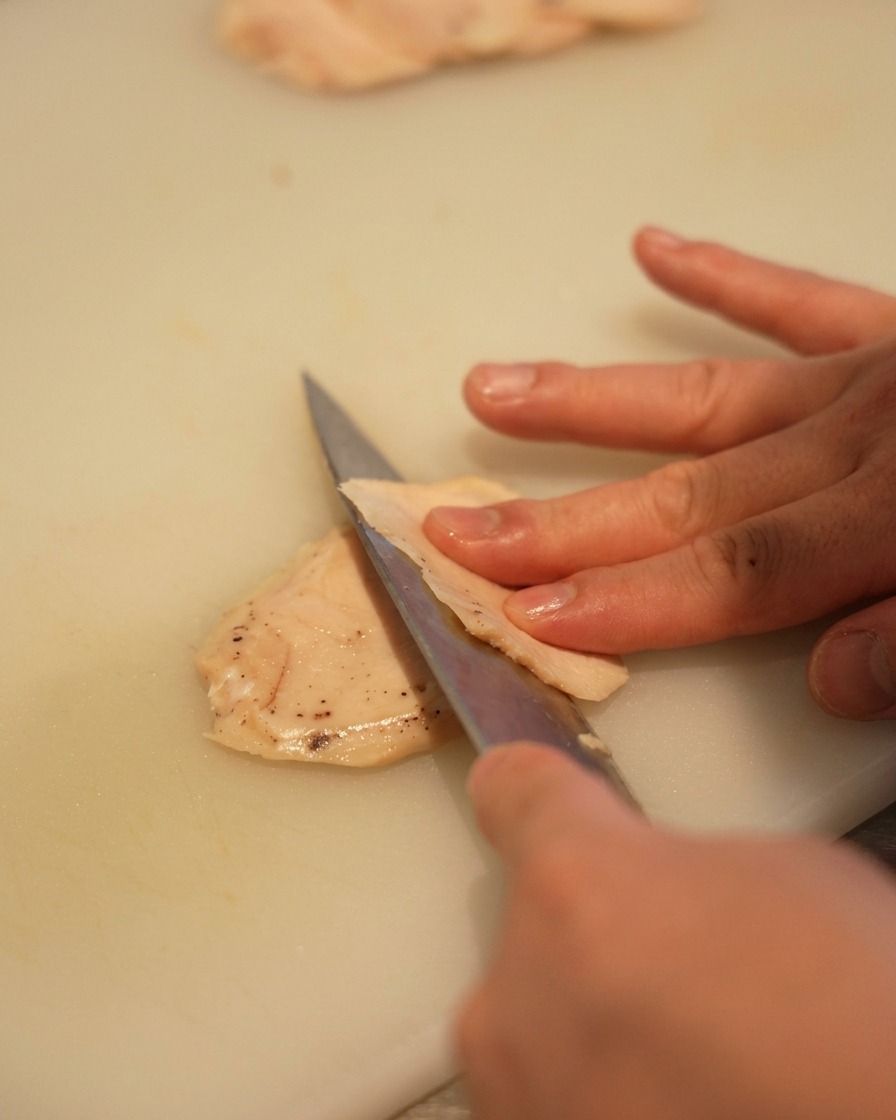 白いまな板の上で、鋭い包丁を使って低温調理した鶏むね肉を薄く切っている様子。