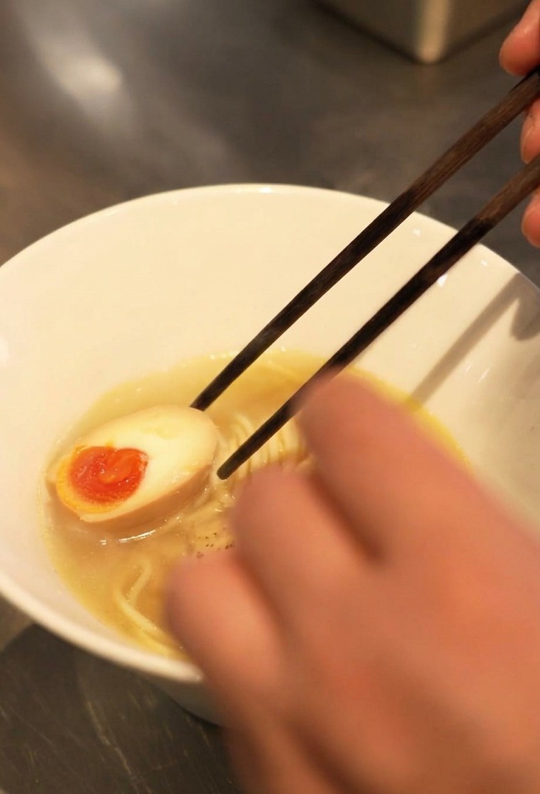 オレンジ色の黄身がとろりとした半熟卵を、ラーメンの丼に添えている様子。