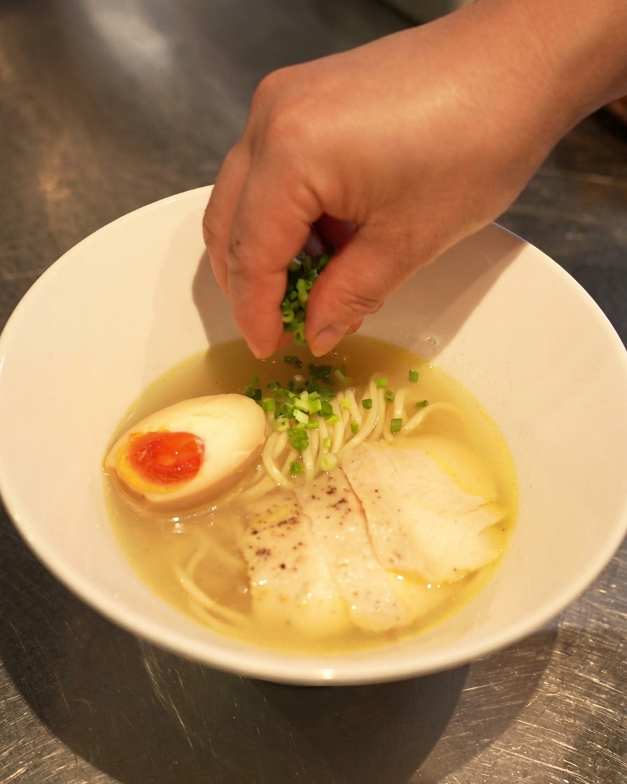 完成した鶏塩ラーメンの上に、手で細かく刻んだねぎを散らしている様子。