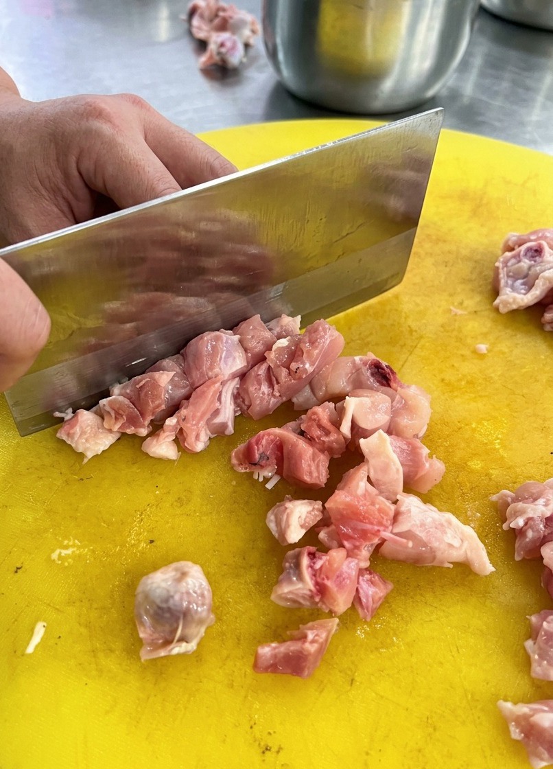 黄色いプラスチックのまな板の上で、中華包丁を使って生の鶏肉を刻んでいる様子。