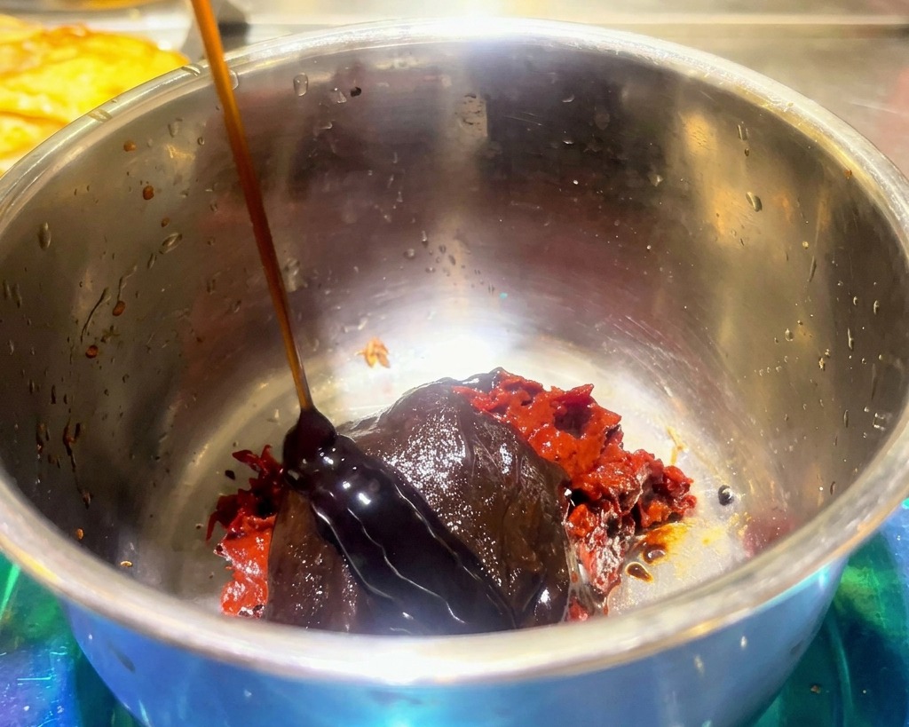 赤い豆板醤や火鍋の素が入ったボウルに、濃い色のソースを注いでいる様子。