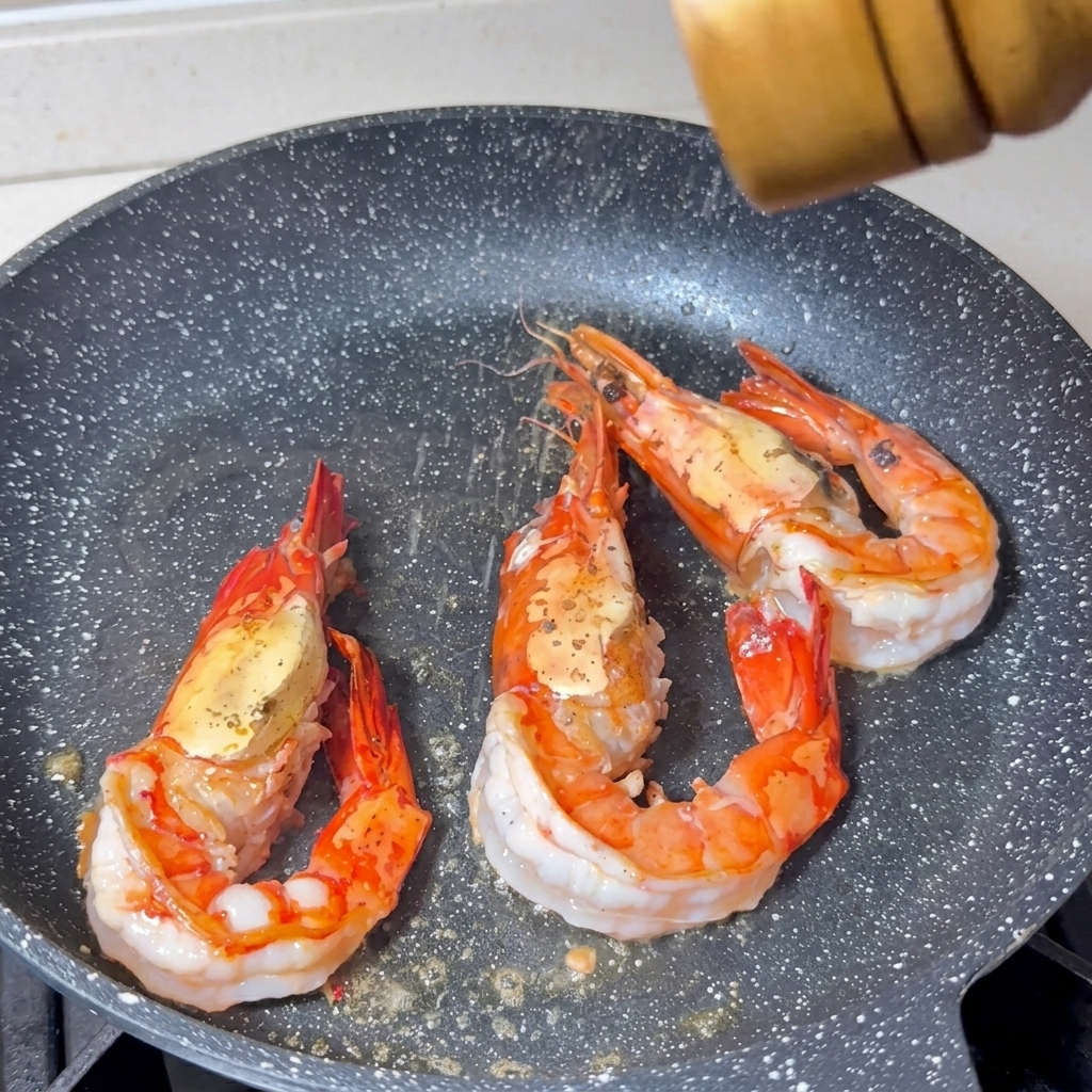 フライパンで焼かれている3尾の大きなエビに塩とこしょうを挽いている様子。