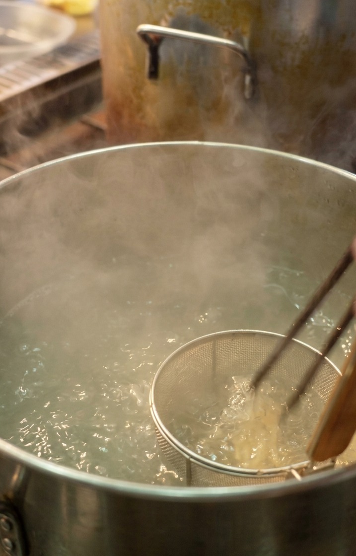 大きな鍋の沸騰したお湯に、テボ（麺揚げザル）を使って細いラーメンの麺を入れる様子。