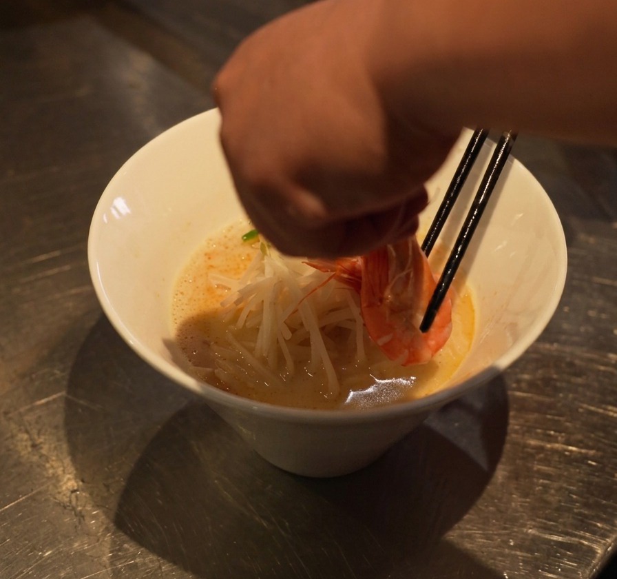 丼の麺ともやしの上に、箸で茹でた海老を丁寧に配置する様子。