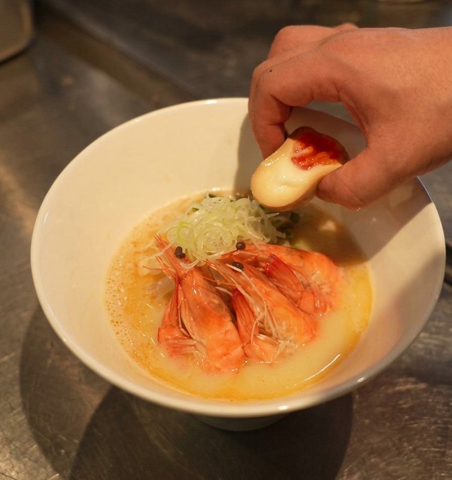 海老やねぎが盛られた丼の脇に、半熟の味付け卵を添える様子。