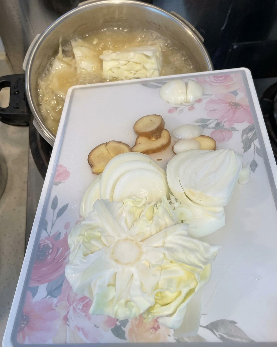 カットした玉ねぎ、生姜、キャベツの芯が乗った白いまな板を傾けて、食材を沸騰した鍋に滑り込ませている様子。