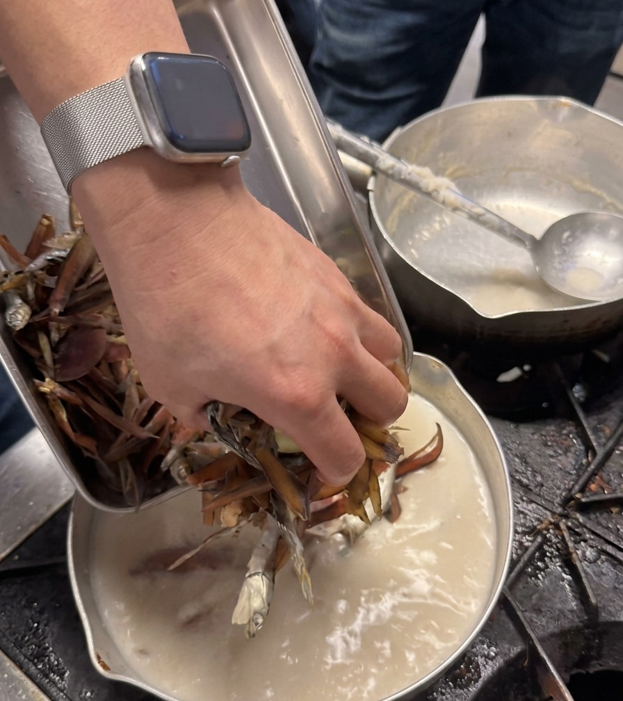 シェフの手が、クリーミーな白いスープが入った鍋の中に、乾燥した濃い茶色の食材をひとつかみ投入している様子。