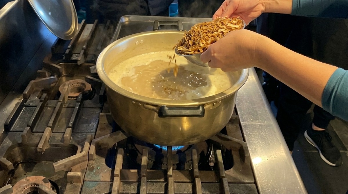 煮立つ白い濃厚スープが入った大きな真鍮の鍋に、手で大量の煮干しを加えている様子。