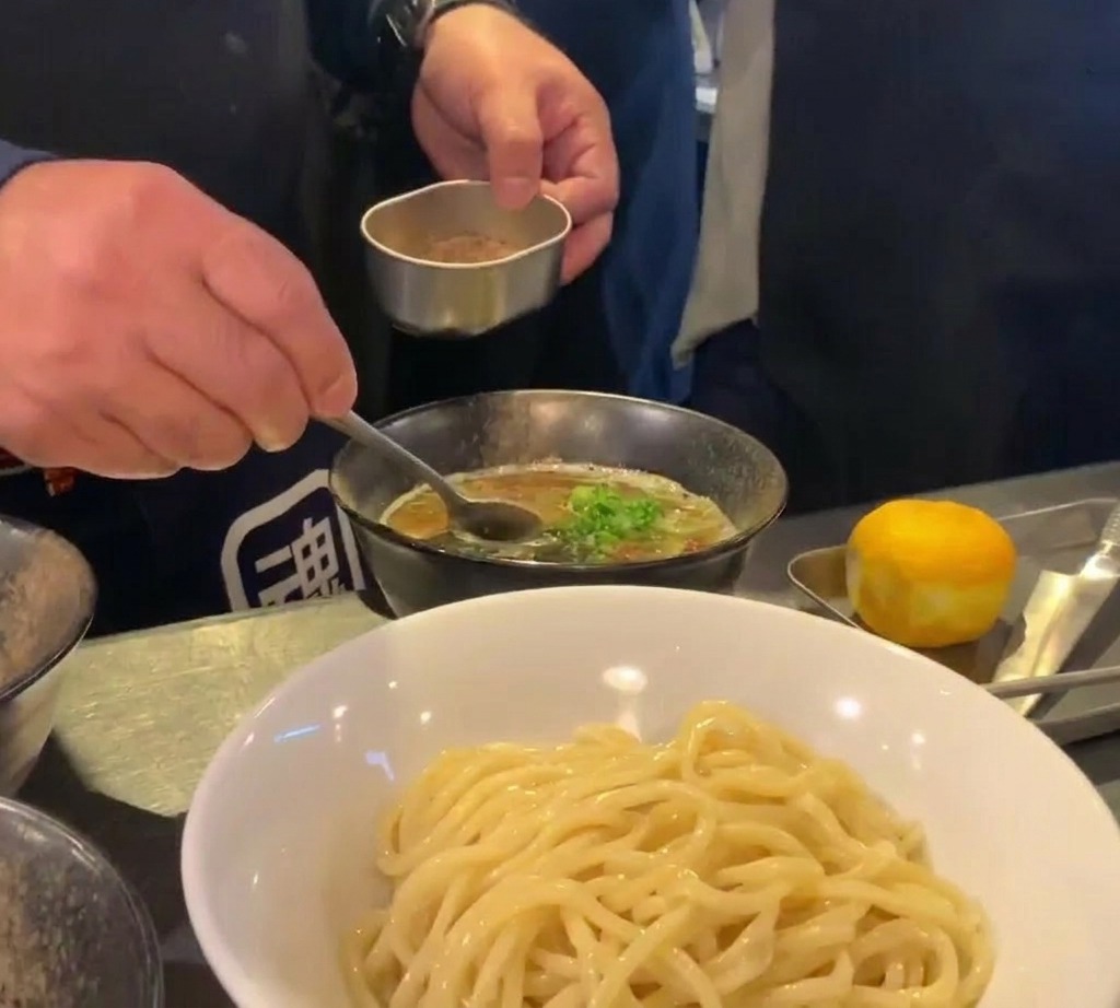 小さな金属容器とスプーンを使い、濃い色の濃厚なつけ汁の上に、きめ細かい茶色の魚粉を振りかける様子。