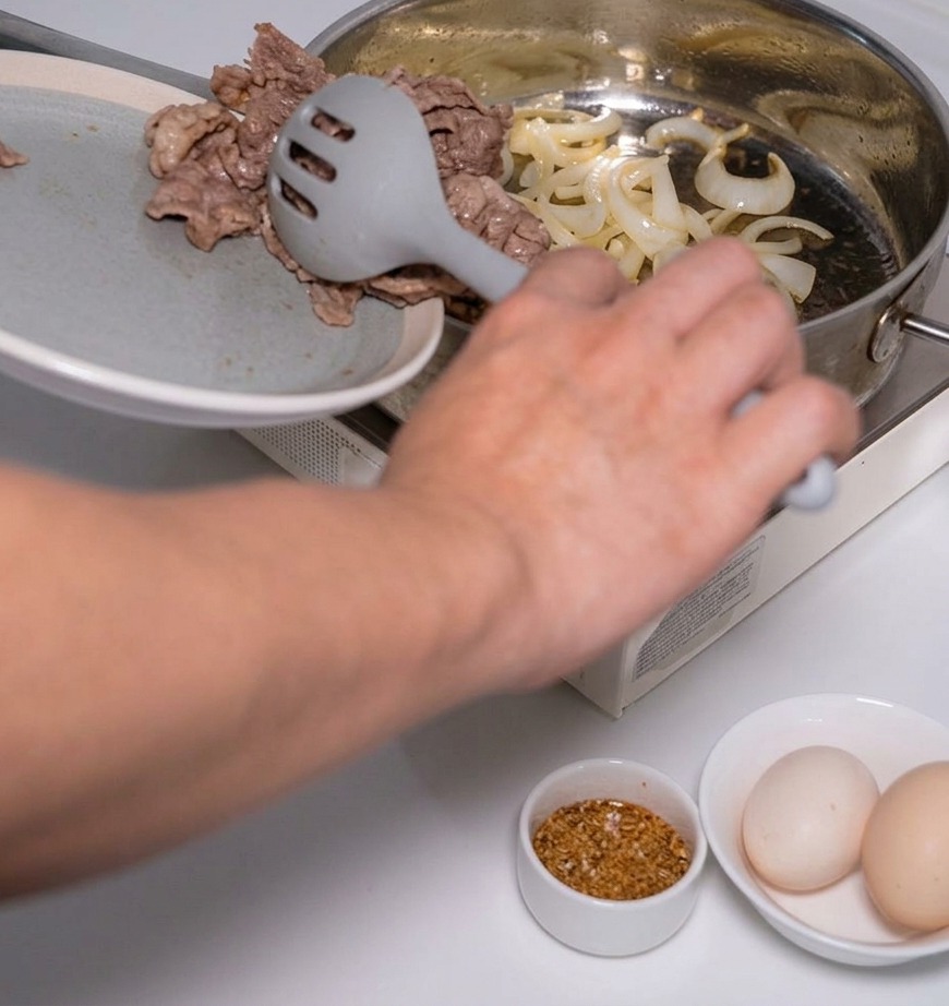 グレーの穴あきスプーンを使い、湯通しした牛肉の薄切りを、玉ねぎを炒めているフライパンに加えている様子。