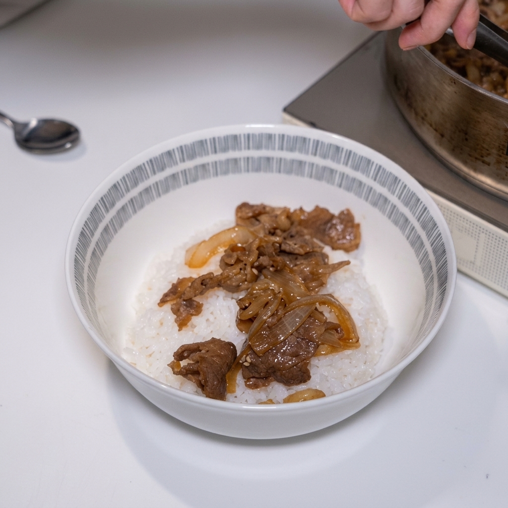 トングを使い、旨味が染み込んだ柔らかい牛肉と玉ねぎを、丼に盛られた温かいご飯の上にたっぷりと盛り付けている様子。